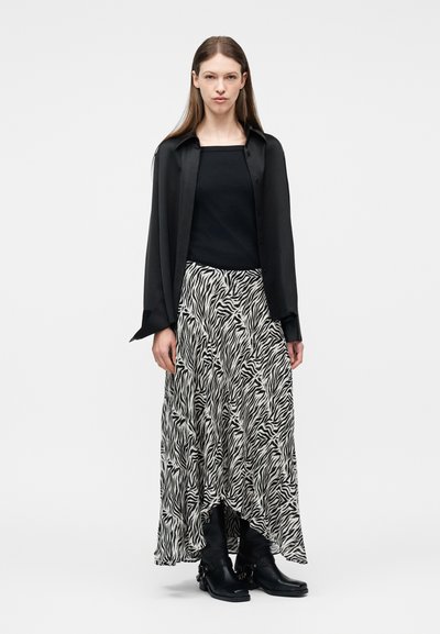 Frau mit langen braunen Haaren, die ein schwarzes Hemd über einem schwarzen Oberteil, einen Rock mit Zebramuster und schwarze Stiefeletten trägt und vor weißem Hintergrund steht.