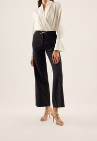 Weiße Wickelbluse mit ausgestellten Ärmeln, kombiniert mit hoch taillierten schwarzen Hosen und goldfarbenen Sandalen mit Absatz und Riemchen.