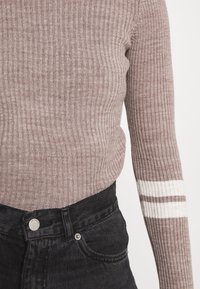 Pull côtelé brun clair avec des rayures blanches près du poignet, associé à un jean en denim noir taille haute.