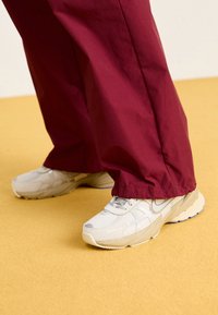 Light grey sneakers with textured surfaces and cushioned soles, paired with loose-fitting burgundy pants, against a yellow background.