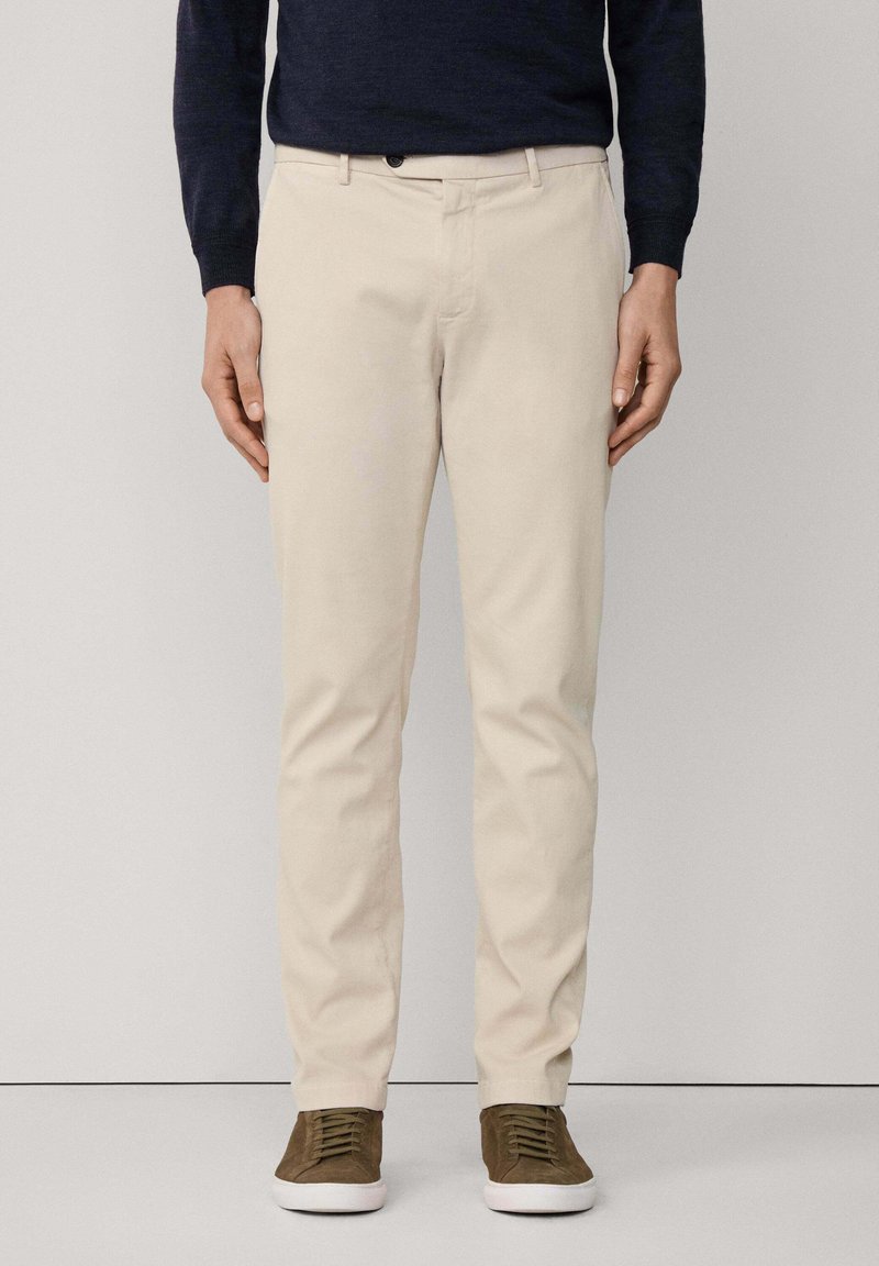 Pantalones de algodón beige con corte recto, que cuentan con bolsillos frontales y un cierre de un botón. Llevados con zapatillas marrones.