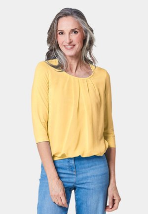 Gelbes Oberteil mit drei Viertelärmeln und gefaltetem Vorderdesign, leichtes Material, runder Ausschnitt, kombiniert mit blauen Jeans.