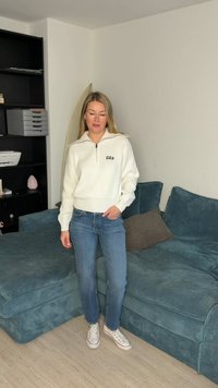 Γυναίκα με λευκή ζακέτα με φερμουάρ και μπλε τζιν στέκεται δίπλα σε μπλε καναπέ από κορδόνι σε σαλόνι.