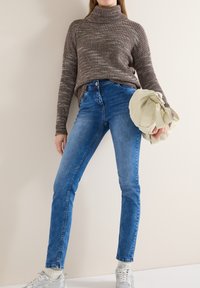 Femme portant un pull à col roulé en maille marron avec un jean bleu et des baskets blanches, tenant une veste beige pliée à la main.