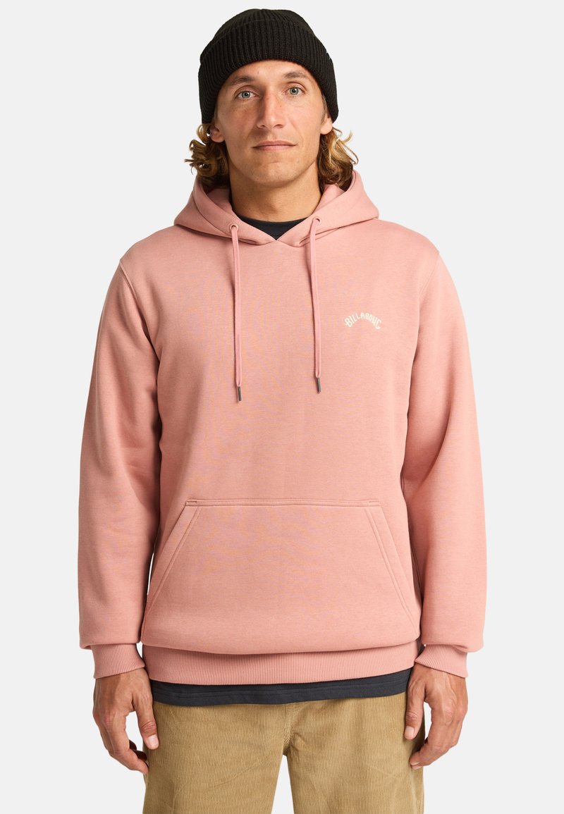 Light pink hoodie made of fleece material, featuring a kangaroo pocket, drawstrings, and a small logo on the chest. Worn over a dark shirt.