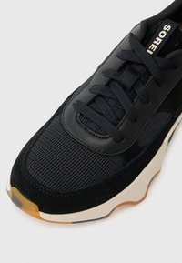 Sorte joggesko med teksturert mesh-overdel, skinn-detaljer og snøring. Sålen er tanfarget med en gjennomsiktig gummidel ved tåen.
