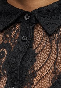 Κοντινό πλάνο γιακά μαύρης δαντέλας με λουλουδένια σχέδια και δύο μαύρα κουμπιά σε διαφανές ύφασμα πάνω στο δέρμα.