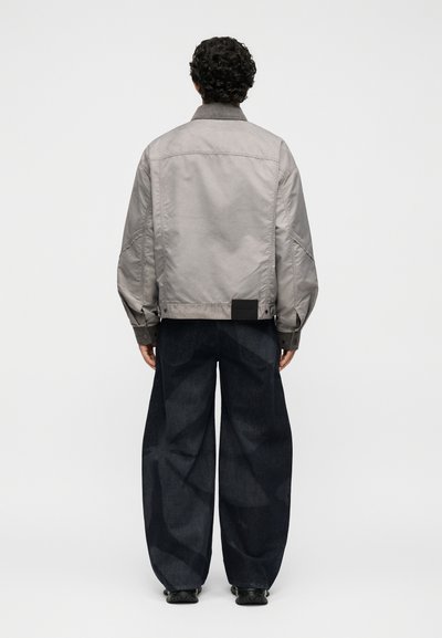 Άτομο με σγουρά μαλλιά που φορά ανοιχτό γκρι μπουφάν και άνετα σκούρα μπλε τζιν, στέκεται στραμμένο σε ένα απλό ανοιχτόφωτο φόντο.