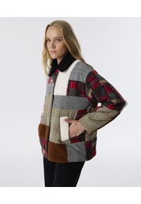 Patchworkjack met gemengde materialen: rode plaid, grijs flanel, crème fleece en bruine nepbont. Bevat twee zakken aan de voorkant en een zwarte kraag.