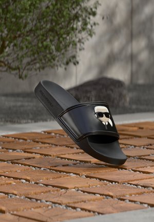 Sort slidesandal med et tegneserieansigt iført solbriller på remmen, vist udendørs på træfliser med grønne planter i baggrunden.