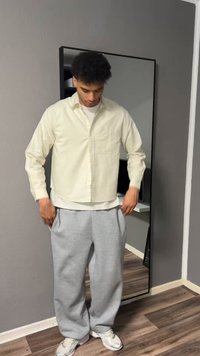 Jeune homme ajustant une chemise beige par-dessus un t-shirt blanc, portant un pantalon de survêtement gris et des baskets blanches, debout devant un miroir en pied à l'intérieur.