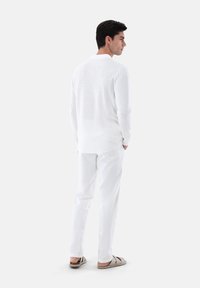 Weißes, gestricktes Langarmshirt mit einem strukturierten Muster, kombiniert mit weißen, eng anliegenden Hosen und hellgrauen Rutschsandalen, von hinten gezeigt.