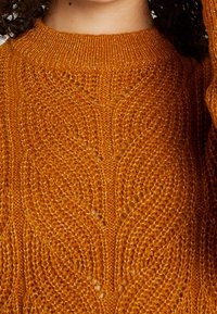Pull orange en maille texturée avec un col côtelé. Présente des motifs ajourés et une coupe ample, mettant en valeur le détail des mailles et la douceur.