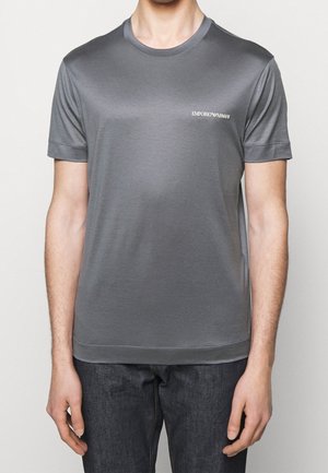 Mand iført en ensfarvet grå Emporio Armani kortærmet t-shirt og mørke jeans, stående med armene afslappet langs siden.