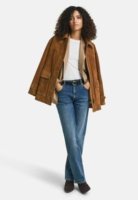 Giacca in suede marrone sopra un maglione beige, abbinata a jeans di denim blu. Il modello sta in piedi con le mani nelle tasche, indossando stivali alla caviglia marroni.