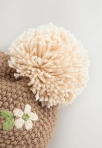 Häkelspielzeug in Braun mit einem großen beigefarbenen Pompon oben, verziert mit gestickten weißen Blumen und grünen Blättern. Weiche, texturierte Oberfläche.