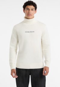 Maglione a collo alto bianco realizzato in tessuto morbido, con un collo e un bordo a coste, e il logo "GUESS JEANS" ricamato in blu scuro.