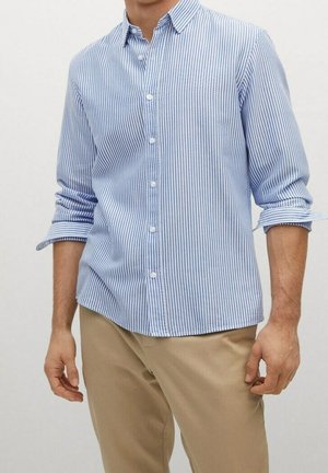 Mężczyzna w jasnoniebiesko-białej koszuli w paski z podwiniętymi rękawami i beżowych spodniach na jednolitym tle.