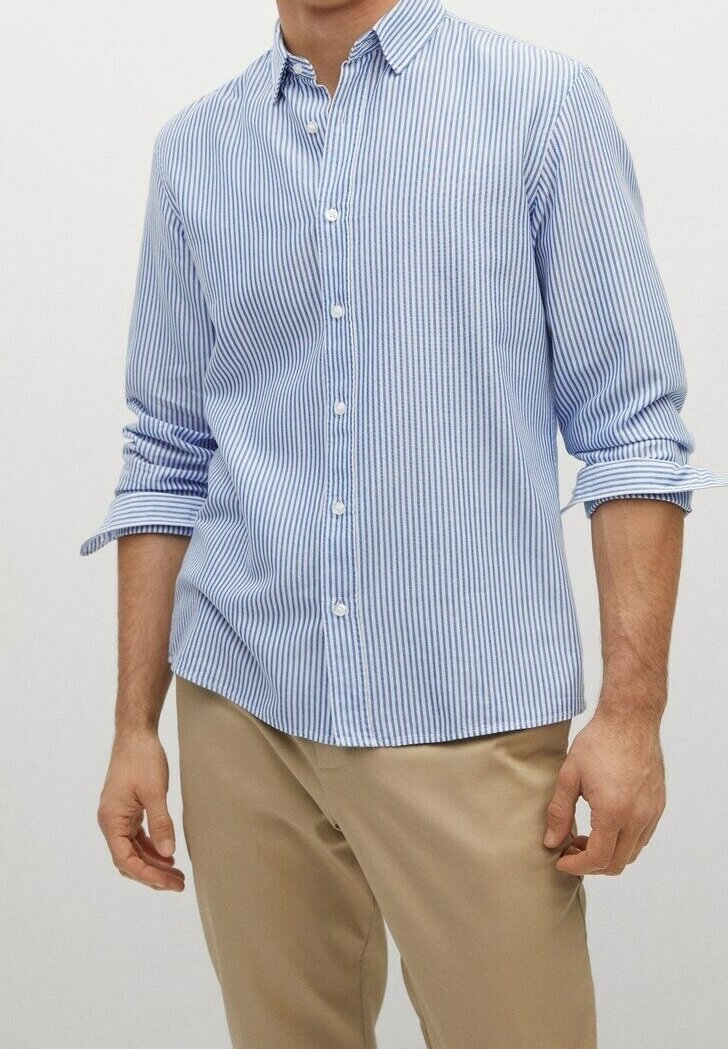 Homme portant une chemise boutonnée à rayures bleu clair et blanches avec les manches retroussées et un pantalon beige, sur un fond uni.