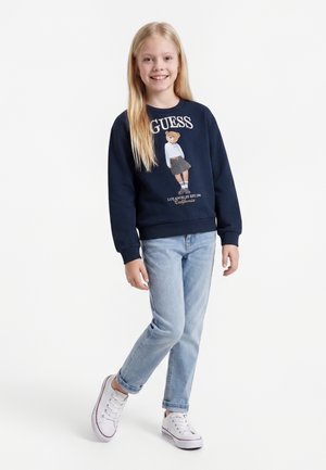 Ragazza sorridente con lunghi capelli biondi che indossa una felpa blu navy con stampa orso, jeans azzurri e scarpe da ginnastica bianche, in piedi su una gamba.