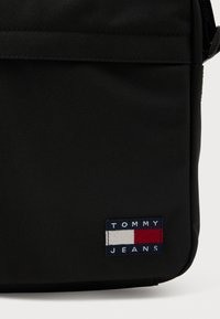 Sort stof skuldertaske med en glat tekstur. Har en rektangulær form og et mærketanvas med blå, røde og hvide vandrette striber.