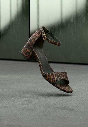 Sandalette mit Leopardenmuster, Blockabsatz und Knöchelriemen mit goldfarbener Schnalle, schwebend über grauem Boden vor reflektierendem grünem Wandhintergrund.