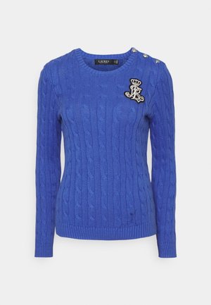 Pull en maille torsadée bleu avec manches longues, trois boutons argentés à l'épaule, et écusson brodé d'une couronne et monogramme sur la poitrine.