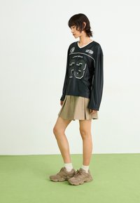 Jersey noir à manches longues avec le numéro de joueur 23 et des accents blancs, associé à une jupe plissée beige et des baskets taupe.