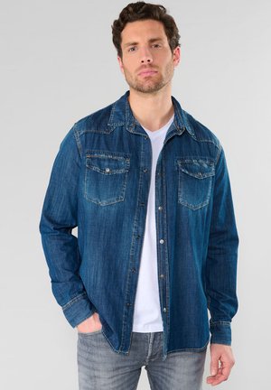 Dunkelblaue Denim-Hemd mit durchgehender Knopfleiste, zwei Brusttaschen und Kragen. Dazu ein weißes T-Shirt darunter.