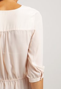 Jasnoróżowa bluzka z okrągłym dekoltem, marszczonym tyłem i podwiniętymi rękawami. Wykonana z gładkiego materiału o delikatnym połysku.