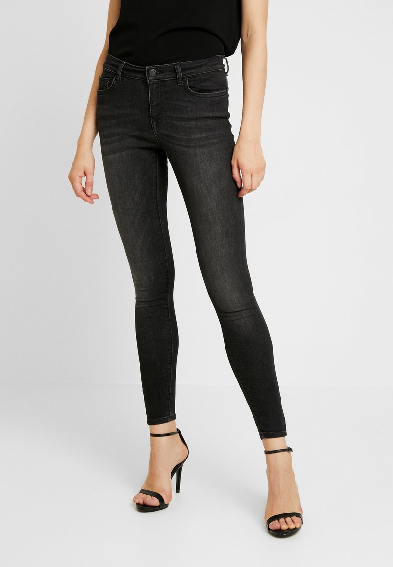 Jean noir slim porté avec un haut noir sans manches et des sandales à talons hauts noires, debout sur un fond clair uni.