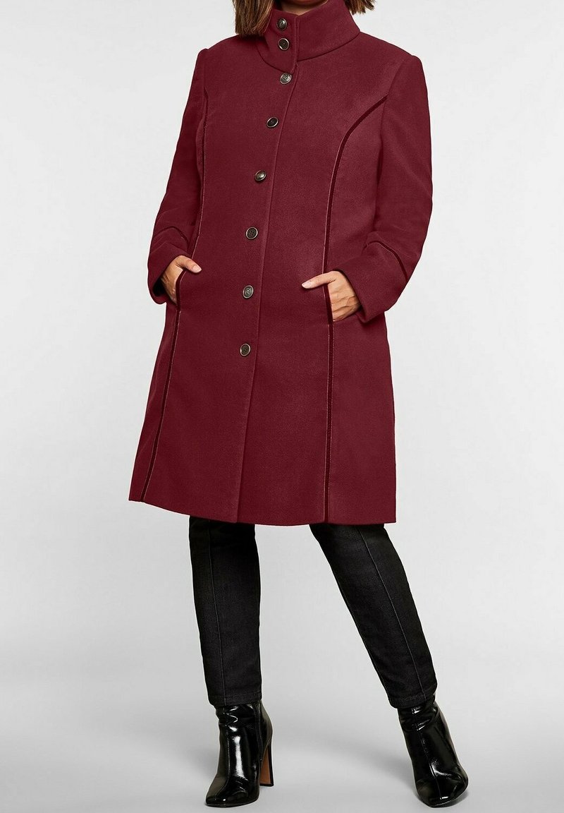 Abrigo de lana burdeos con cuello alto, cierre frontal de botones prominente, bolsillos laterales y acentos de costura contrastantes, combinado con pantalones negros y botas.