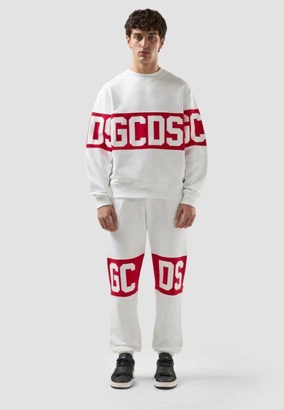 Ung man som bär en vit sweatshirt och byxor med kraftiga röda band och vita blockbokstäver "GCDS" över bröst och knän, svarta sneakers.