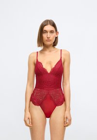 Rød blonde bodysuit med justerbare stropper, dyp V-hals, intrikat blomsterdesign og gjennomsiktig nedre panel som fremhever hoftene.