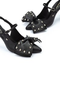 Tacones slingback de cuero negro con punta afilada, adornados con un gran lazo y tachuelas doradas, con una delgada correa en el tobillo y un acabado texturizado.