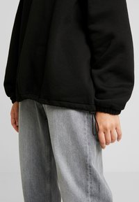 Sudadera negra con dobladillo y puños elásticos, con un ajuste holgado. Combinada con vaqueros gris claro de corte ancho. Mano apoyada al costado.