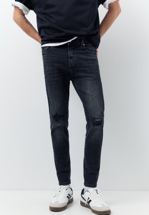 Bărbat îmbrăcat în blugi skinny negri, rupți, adidași albi cu dungi negre și un tricou negru cu mânecile suflecate.