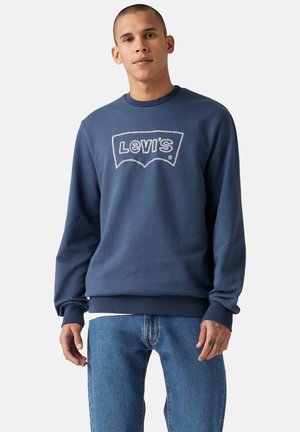 Granatowy bawełnowy sweatshirt z białym haftowanym logiem "Levi's". Posiada okrągły dekolt, ściągacze przy mankietach oraz luźny krój. Tło w denimu.