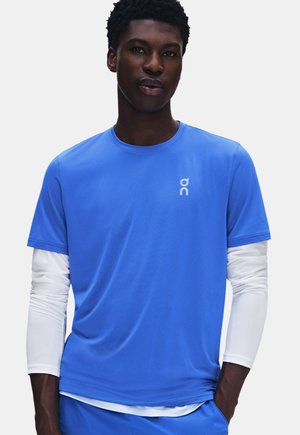 Homme portant une chemise bleue vif à manches courtes par-dessus une chemise blanche à manches longues, debout avec une main dans la poche devant un fond uni.