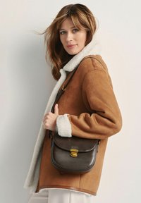 Brązowy zamszowy płaszcz z białym kołnierzem i mankietami z owczej wełny, w połączeniu z małą ciemnobrązową skórzaną torbą na ramię z złotym zapięciem.