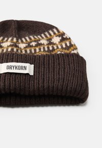 Barna kötött sapka mintás tetejével bézs és sárga színben. Bordás szegéllyel rendelkezik, és egy kis fehér címke található rajta fekete felirattal, ami "DRYKORN".