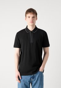 Polo shirt nera con colletto a zip, caratterizzata da un bordo grigio e un logo discreto sul petto. Abbinata a jeans in denim azzurro chiaro.