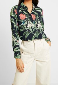 Blusa floreale in tessuto blu scuro, con fiori colorati e foglie verdi, chiusura frontale con bottoni, maniche lunghe e colletto classico.