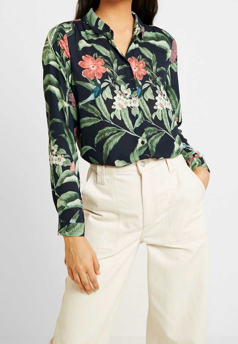 Blusa floreale in tessuto blu scuro, con fiori colorati e foglie verdi, chiusura frontale con bottoni, maniche lunghe e colletto classico.