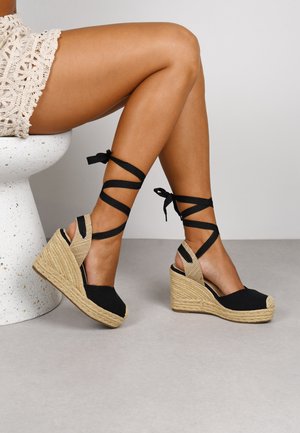 Black espadrille wedge sandals with braided jute sole, open toe, and crisscross black ribbon straps wrapping around the ankle.