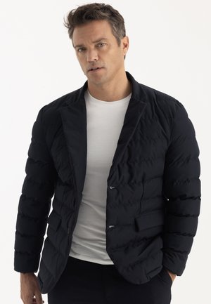 Homme portant un blazer matelassé noir sur un t-shirt blanc, debout devant un fond clair uni avec une main dans la poche.