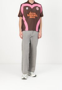 Brun sportströja med rosa accenter och orange text, kombinerad med ljusgrå vida jeans och grå sneakers. Enkelt kragsdesign.