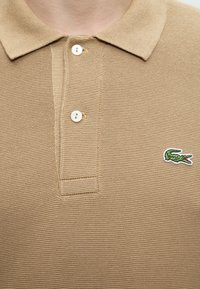 Polo beige texturé avec deux boutons blancs et un petit logo crocodile vert sur la poitrine gauche.