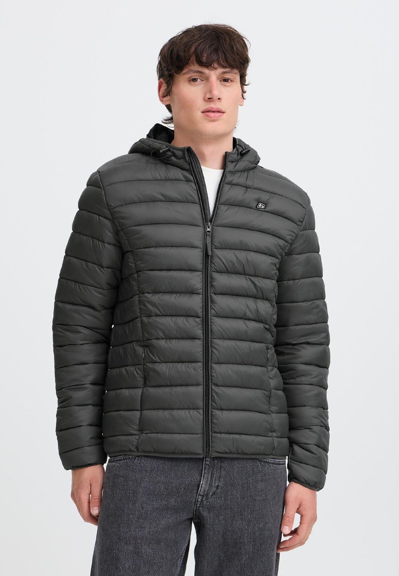Joven con chaqueta negra acolchada con capucha y cremallera y vaqueros gris oscuro, de pie frente a un fondo claro y liso.