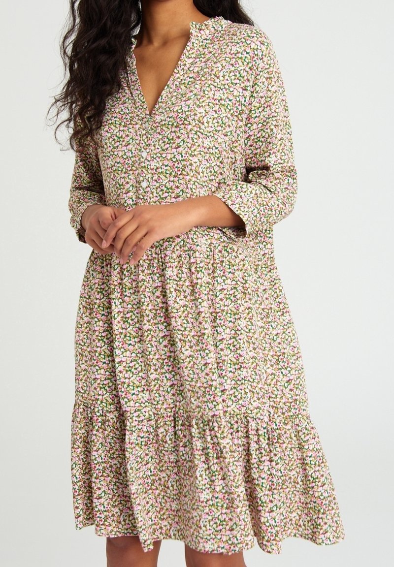 Femme portant une robe mi-longue à manches longues avec un motif floral rose, blanc et vert et un détail de jupe froncée.